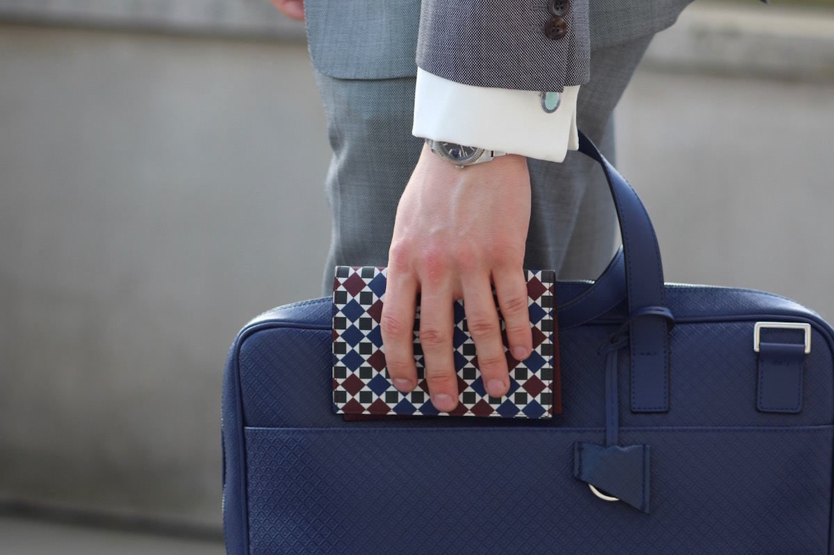 A stylish man in a suit carrying the Oxford Laptop Bag and City Cards Holder from MARK / GIUSTI, walking through the financial district. Featured in The Rake Magazine collaboration, showcasing a dapper and rakish look.