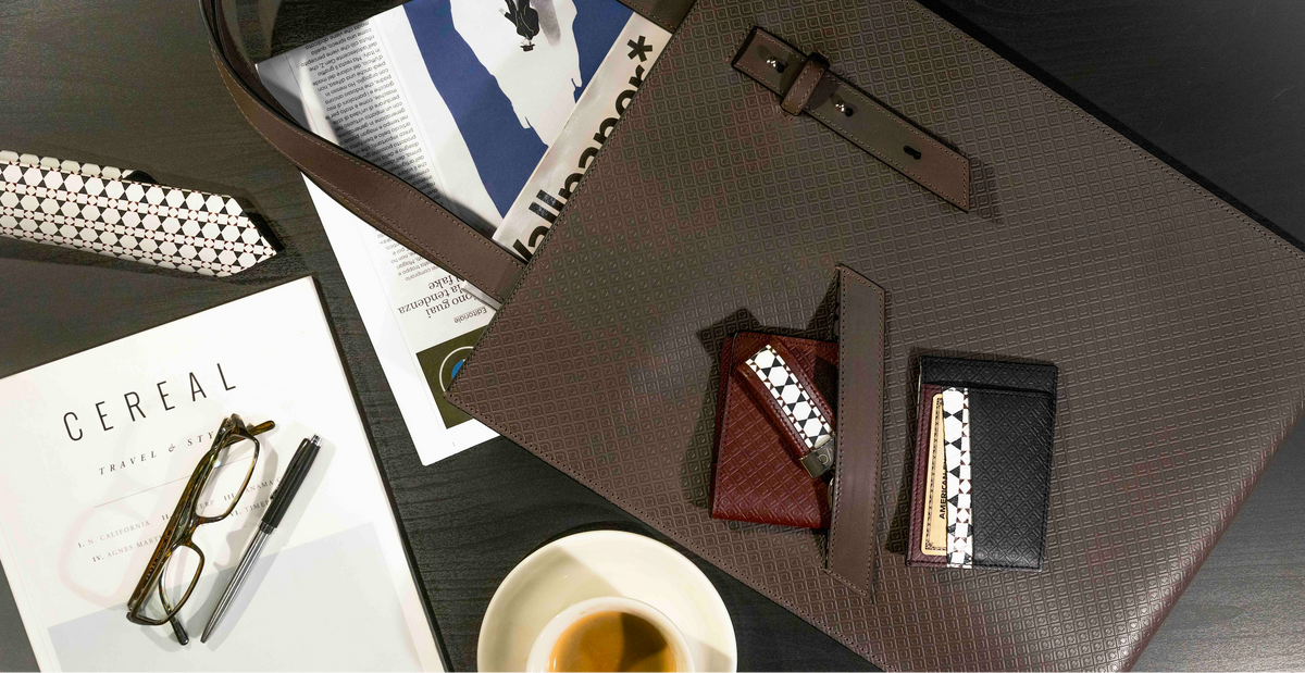 MARK / GIUSTI handcrafted in italy leather Firenze business bag with the Heathrow passport wallet and bank cards holder laid open on a dark tabletop, surrounded by accessories including mosaic patterned leather tie, a magazine, eyeglasses, a pen, and a cup of coffee
