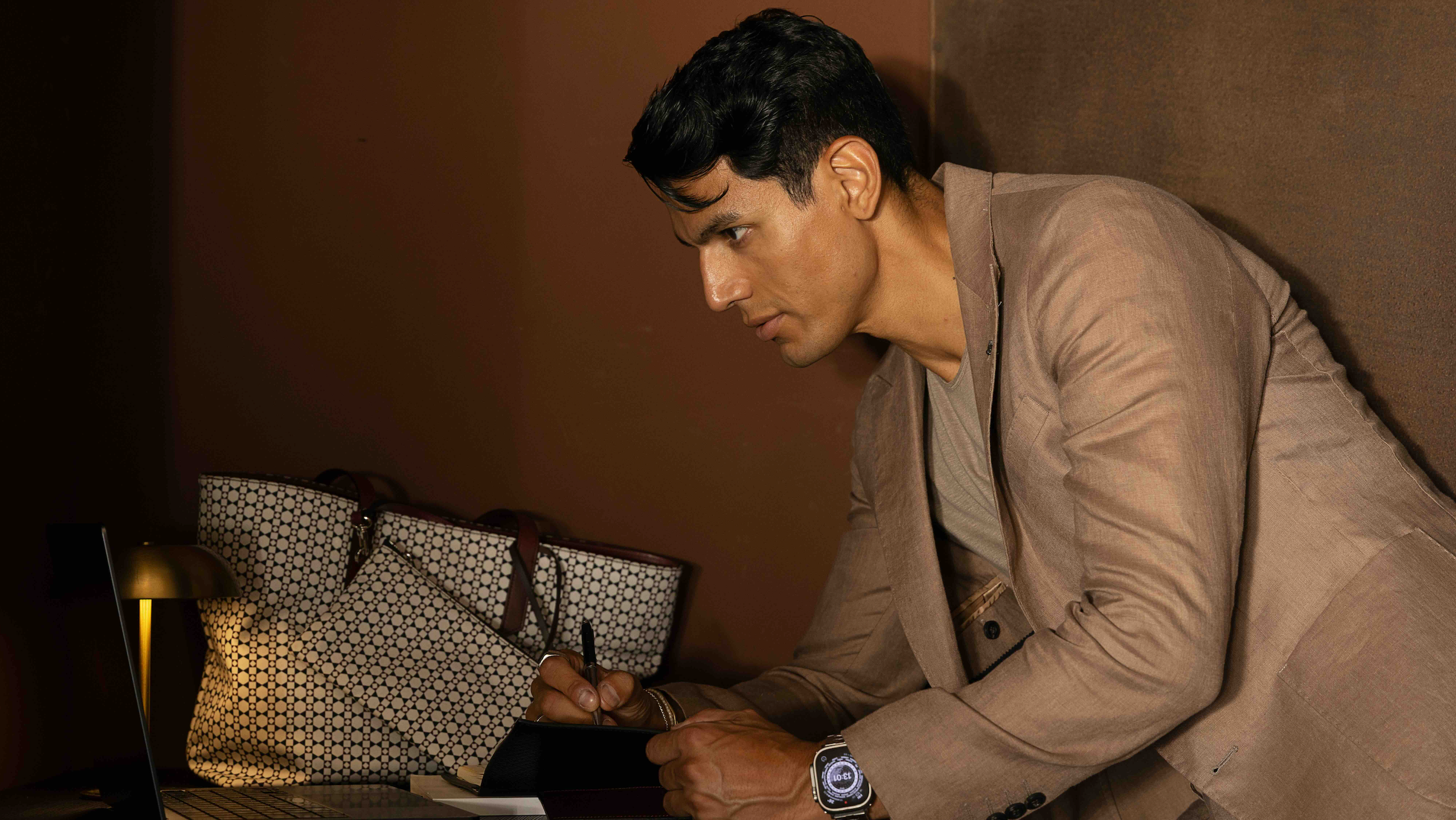 Brand Ambassador Cristiano D'angelo stars in the latest MARK / GIUSTI movie "Fragments of a Day". Here sitting at a desk with a laptop and mosaic Ischia Tote bag, wearing a taupe suit.