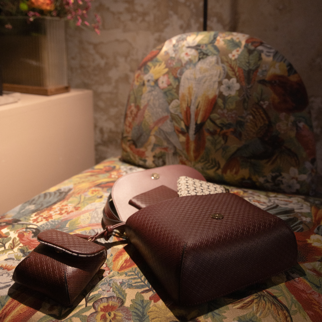A MARK / GIUSTI  handcrafted bellagio handbag set with matching accessories, including a small museo wallet and Heathrow passport wallet and a zippered case, arranged on a floral-patterned upholstered chair in a warm indoor setting, part of the Milan Design Week Collaboration with Youssef El Hadi.