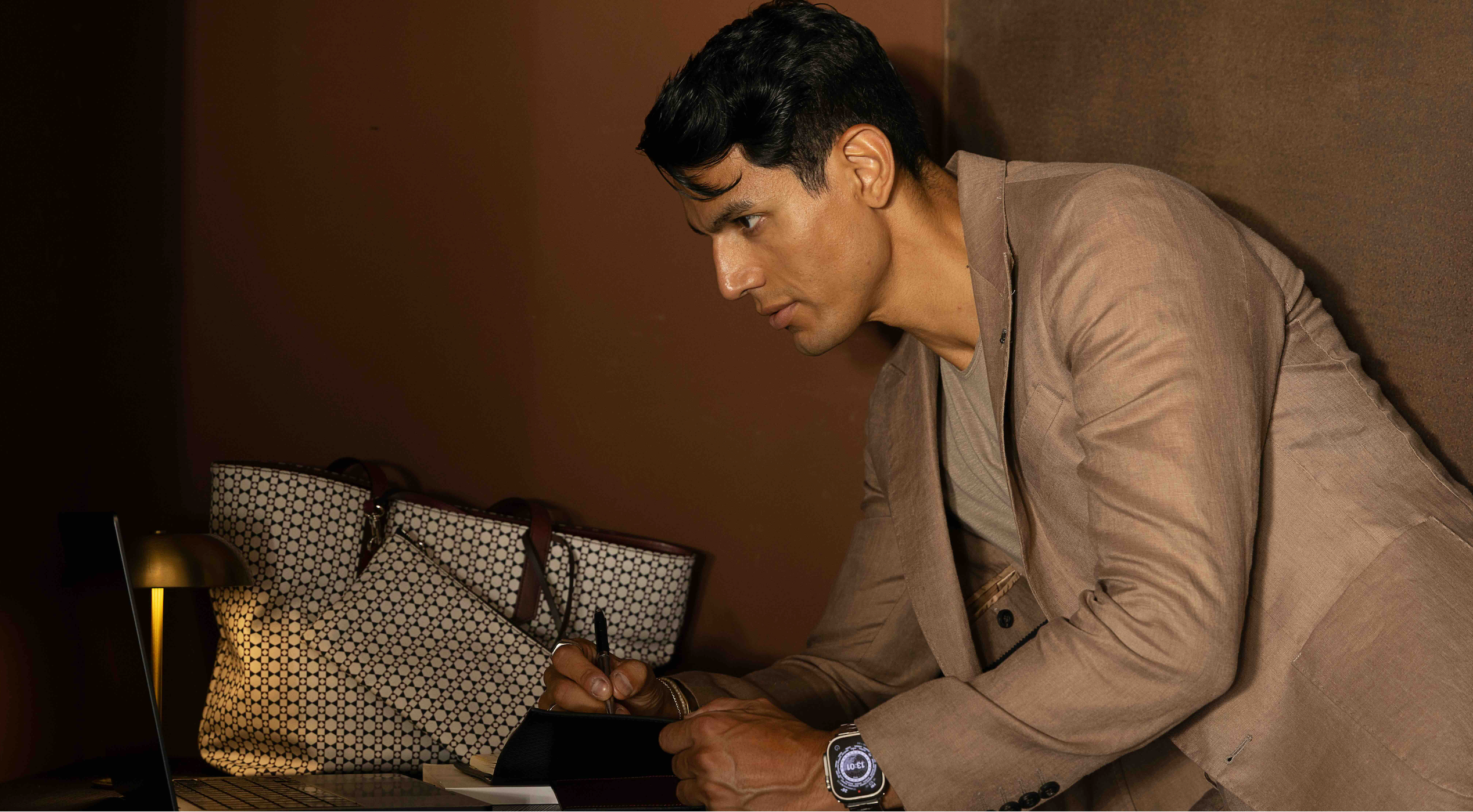 Brand Ambassador Cristiano D'angelo stars in the latest MARK / GIUSTI movie "Fragments of a Day". Here sitting at a desk with a laptop and mosaic Ischia Tote bag, wearing a taupe suit.